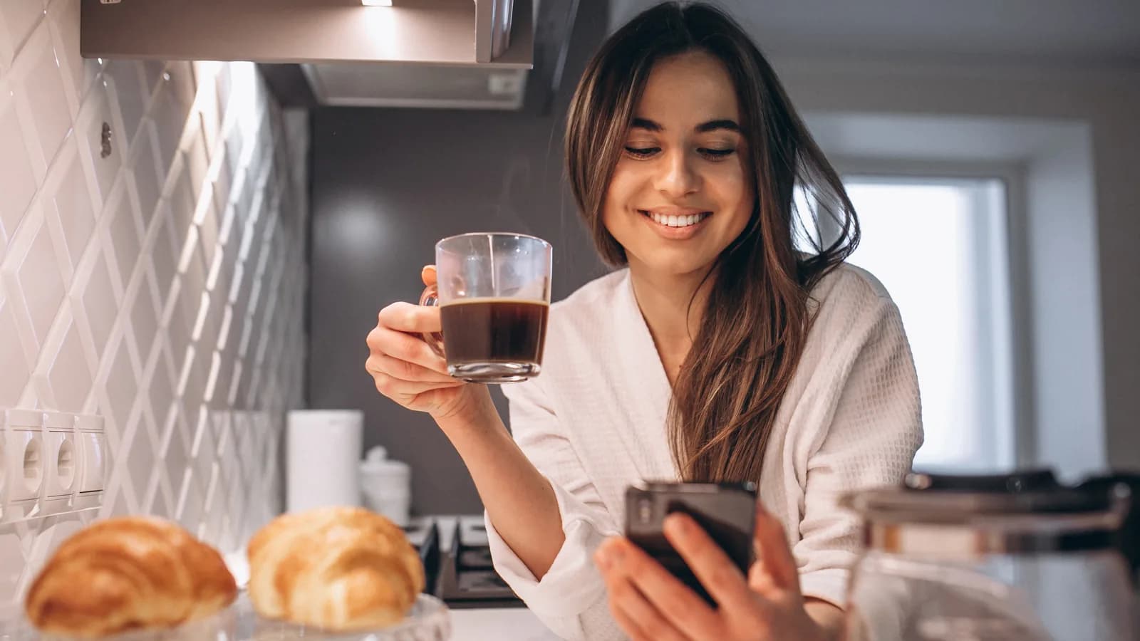 Une femme lit ses e-mails sur son téléphone, une tasse de café à la main, dans sa cuisine